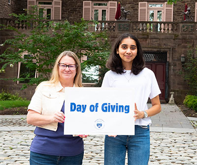 Two Bucks students pose on a cobblestone walkway in front of Tyler Hall, holding a “Day of Giving” sign.