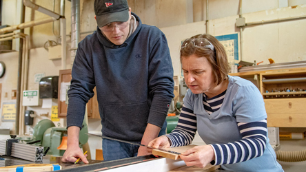 Female measures piece of wood with male standing at side