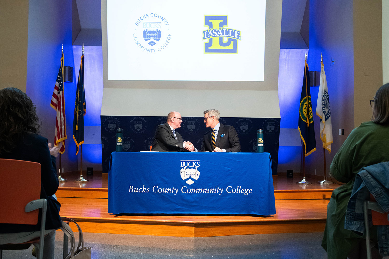 Bucks County Community College President & CEO Patrick M. Jones, Ph.D. and La Salle University President Daniel J. Allen, Ph.D. sit at table with a blue Bucks County Community College cloth, shaking hands under projected logos.