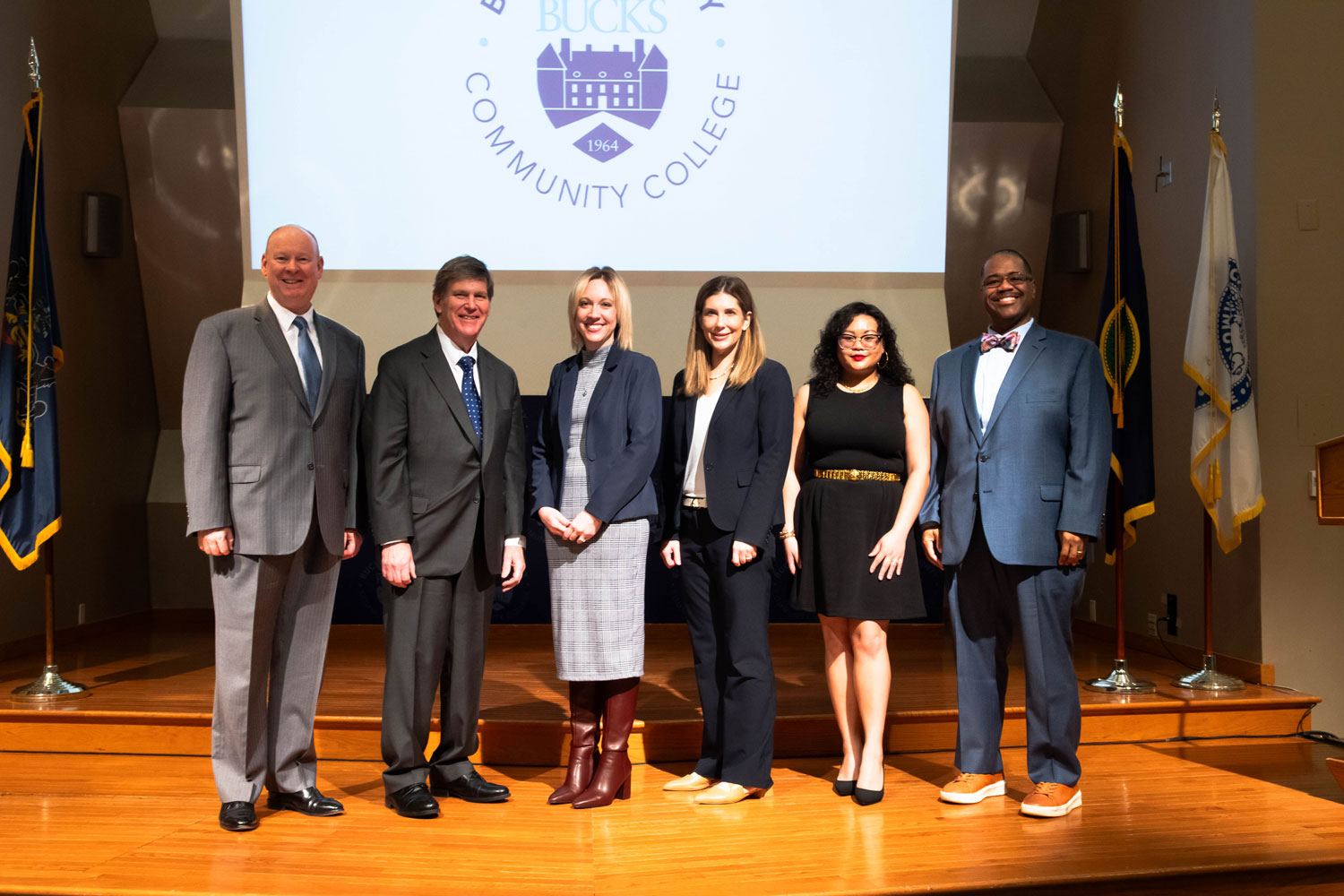 Bucks County Community College president and staff stand on a wooden stage with a Pennsylvania Department of Education representative, all in professional attire and smiling. A Bucks County Community College logo is projected on a screen behind them.