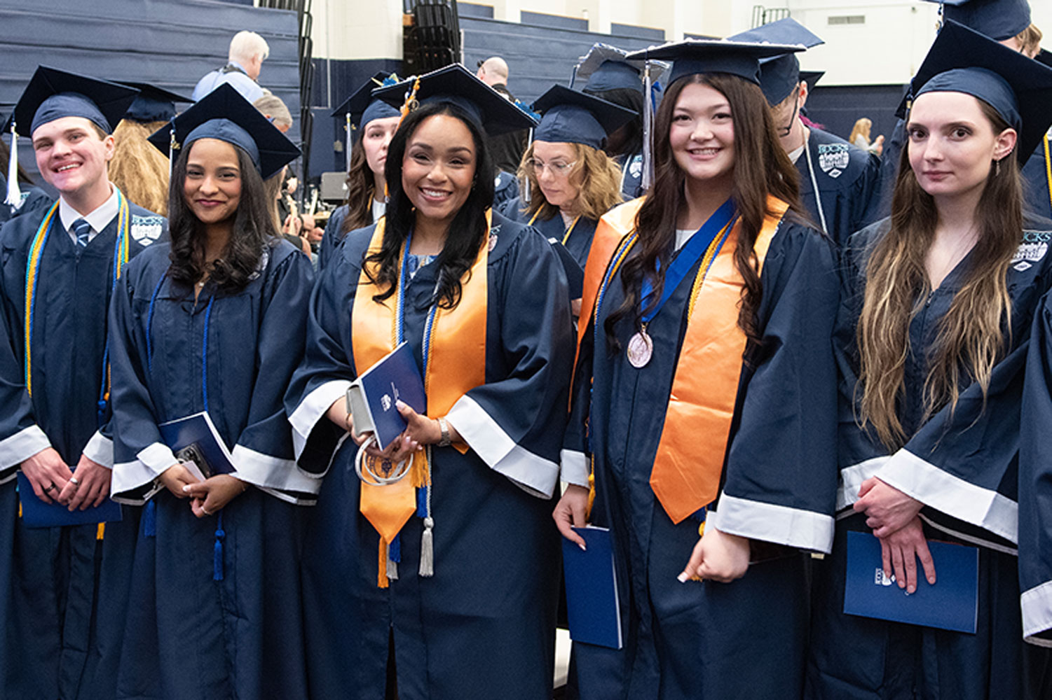 A diverse group of graduates in blue gowns and caps smile proudly. Two wear orange stoles. They hold diplomas, celebrating in a cheerful mood.