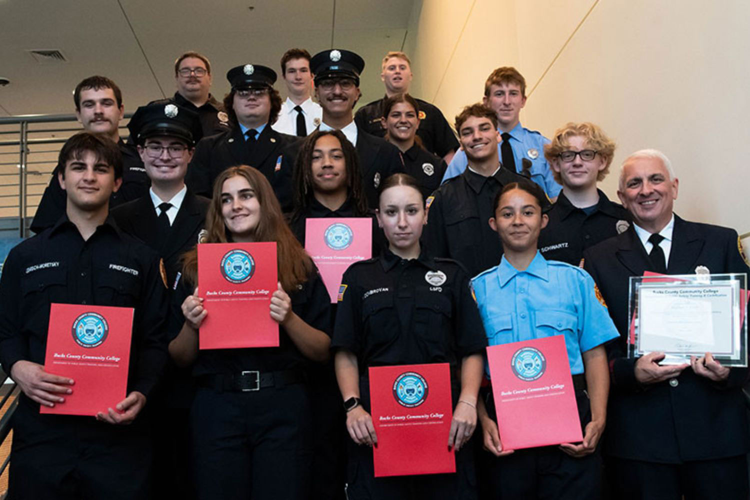 A diverse group of students and instructors in public service uniforms poses for a photo, holding certificates in a bright indoor setting, commemorating a community college graduation or ceremony.