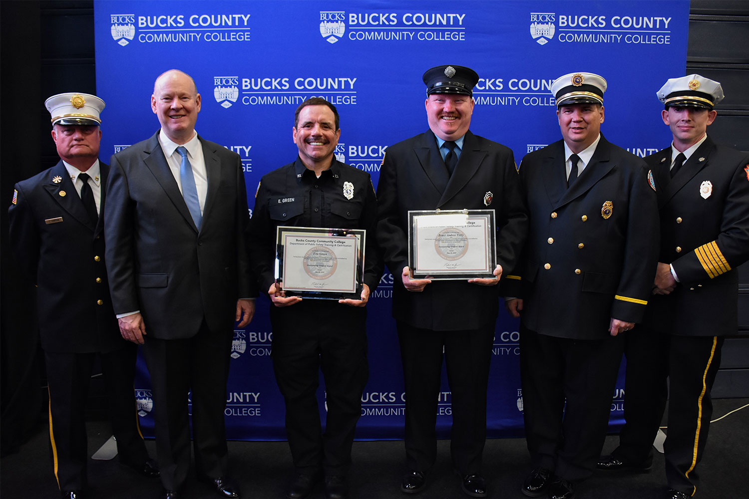 Six men pose in front of a Bucks County Community College backdrop, with two holding framed certificates.