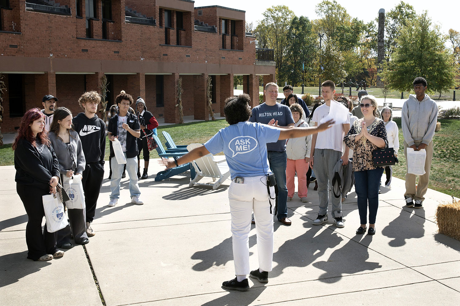 A group gathers outside a brick building while a person in an “ASK ME!” shirt gestures toward them.