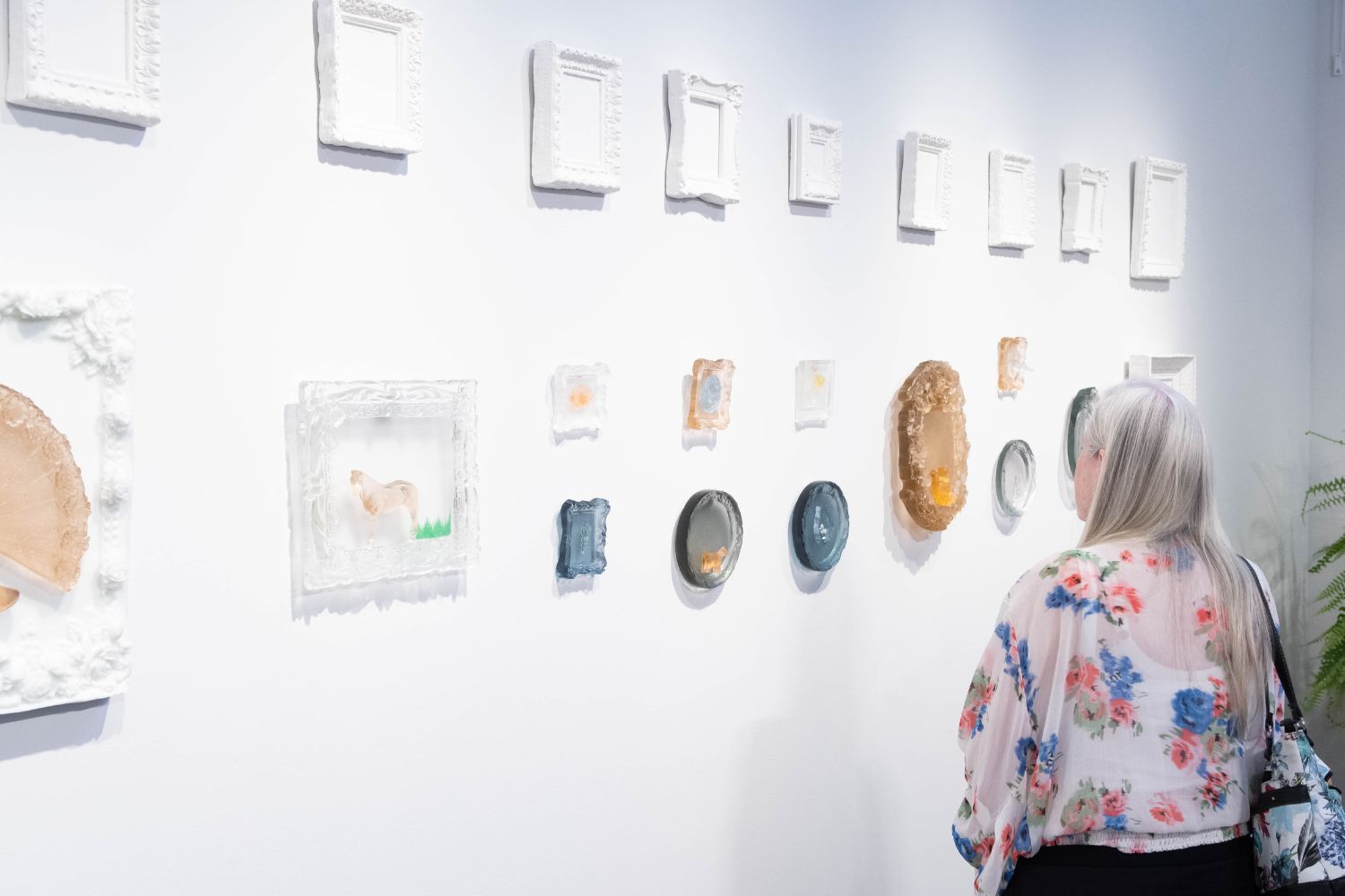 Person viewing an arrangement of ornate white frames and colorful sculptural artworks on a gallery wall.