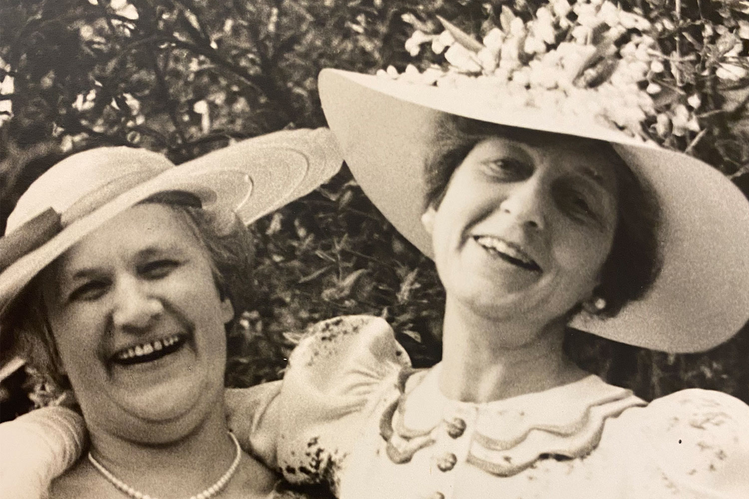 Sepia photograph of two smiling women in wide-brimmed hats posing outdoors among foliage.